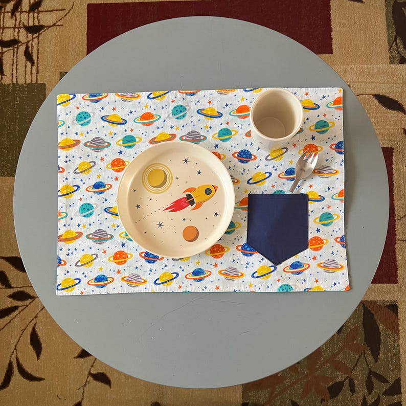 May include: A white placemat with a space-themed pattern featuring planets, stars, and a rocket ship. A white plate with a similar design is placed on top of the placemat, along with a blue napkin holder and a white cup.