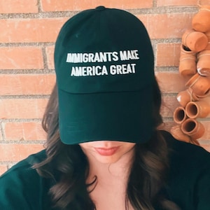 May include: A dark green baseball cap with the text "IMMIGRANTS MAKE AMERICA GREAT" in white block letters. The cap is worn by a person with long, dark hair.