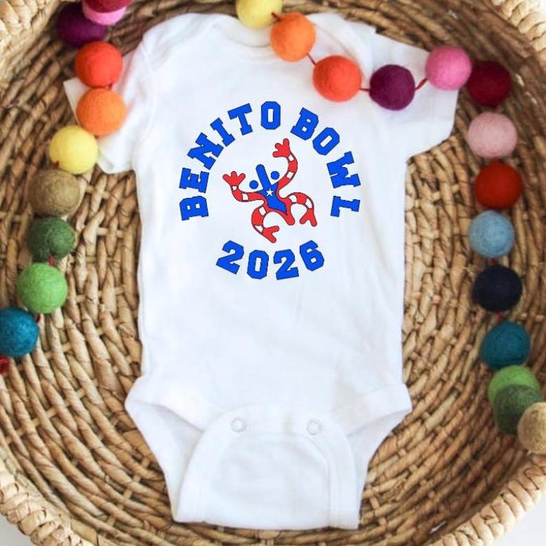 Op de afbeelding: Witte babybody met de blauwe tekst "BENITO BOWL 2026". De body heeft een rood, wit en blauw ontwerp. Gepresenteerd op een rieten mand met een slinger van kleurrijke vilten ballen.