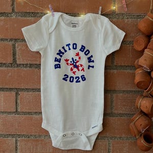 Op de afbeelding: Witte babybody met de tekst "BENITO BOWL 2026" in blauw, met een rood-witte afbeelding. De body hangt aan een waslijn met knijpers tegen een bakstenen muur.