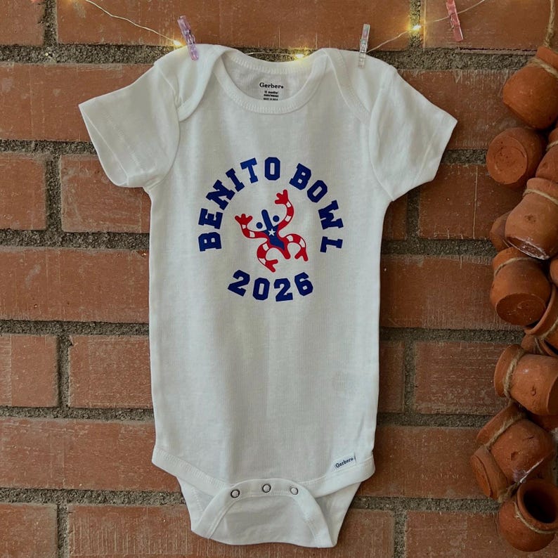 Op de afbeelding: Witte babybody met de tekst "BENITO BOWL 2026" in blauw, met een rood-witte afbeelding. De body hangt aan een waslijn met knijpers tegen een bakstenen muur.
