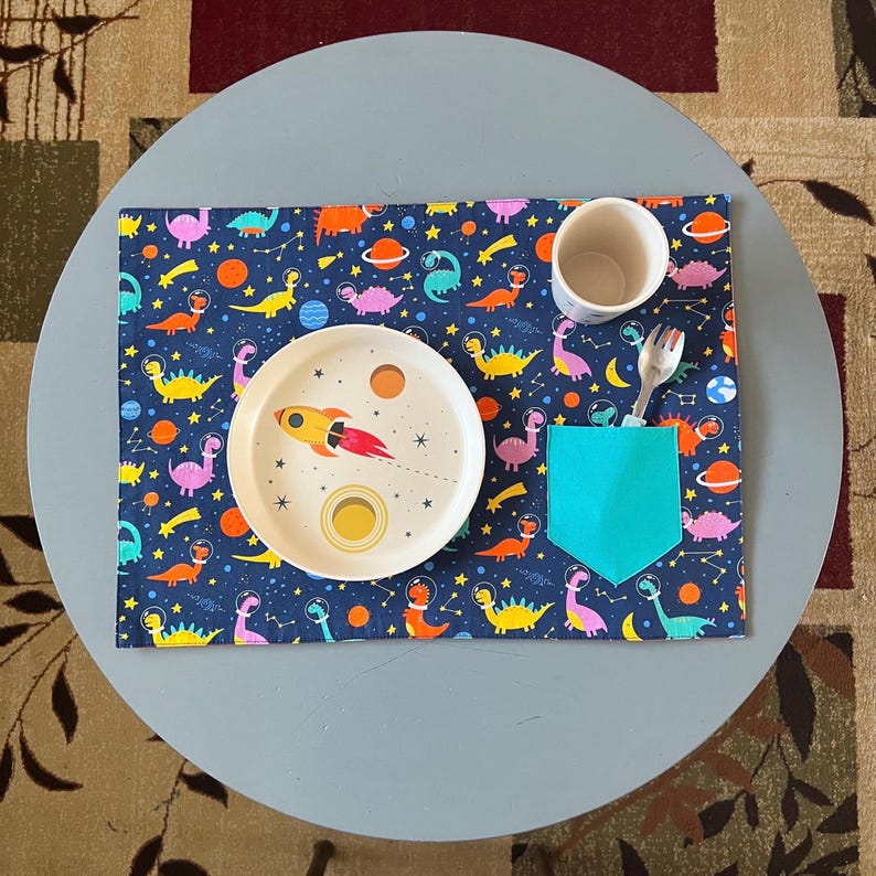 May include: A blue placemat with a dinosaur and rocket ship pattern. A white plate with a rocket ship and planets design, a blue cup, and a silver fork are on the placemat.