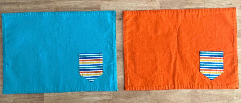 May include: Two placemats, one turquoise blue and one orange, each with a colorful striped pocket sewn on.