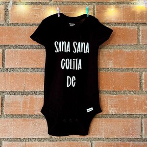 May include: Black baby onesie with white text that reads "Sana Sana Colita De".