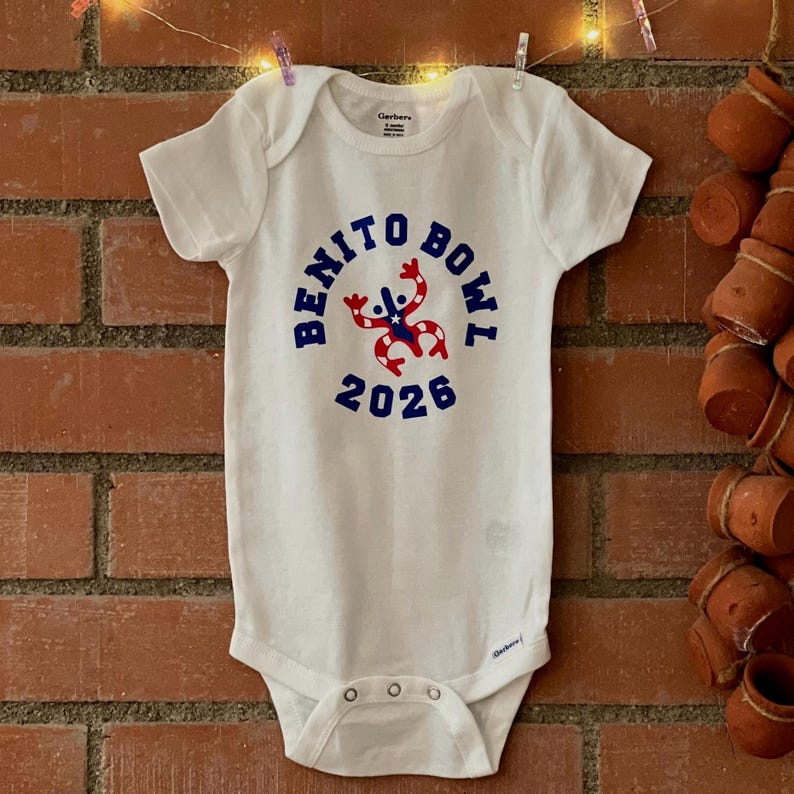 Op de afbeelding: Witte babybody met de tekst "BENITO BOWL 2026" in blauw en rood. De body heeft korte mouwen en drukkers aan de onderkant. Het ontwerp bevat een cartoonfiguur.