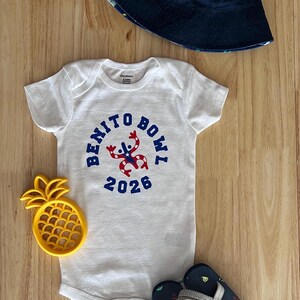 Op de afbeelding: Een witte babybody met de tekst "BENITO BOWL 2026" in blauw, een marineblauwe vissershoed, een geel ananasvormig speelgoed en marineblauwe teenslippers met nautische ontwerpen. Op een houten oppervlak.