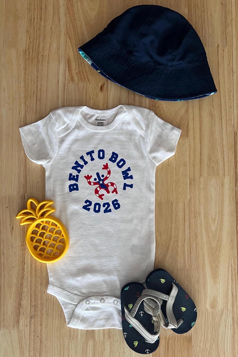 Op de afbeelding: Een witte babybody met de tekst "BENITO BOWL 2026" in blauw, een marineblauwe vissershoed, een geel ananasvormig speelgoed en marineblauwe teenslippers met nautische ontwerpen. Op een houten oppervlak.