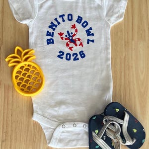 Op de afbeelding: Witte babybody met de tekst "BENITO BOWL 2026" in blauw, met een rood-witte afbeelding. Een gele ananasvormige koekjessteker en marineblauwe teenslippers met witte bandjes en ankerontwerpen zijn ook aanwezig.