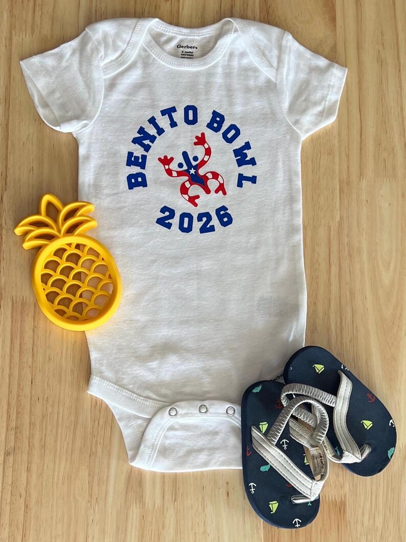 Op de afbeelding: Witte babybody met de tekst "BENITO BOWL 2026" in blauw, met een rood-witte afbeelding. Een gele ananasvormige koekjessteker en marineblauwe teenslippers met witte bandjes en ankerontwerpen zijn ook aanwezig.
