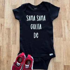 May include: Black baby onesie with white text that reads "Sana Sana Colita De". The onesie is laying on a wooden surface with a pair of red Vans shoes and blue sunglasses.