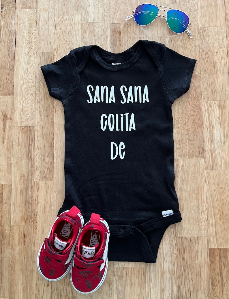May include: Black baby onesie with white text that reads "Sana Sana Colita De". The onesie is laying on a wooden surface with a pair of red Vans shoes and blue sunglasses.