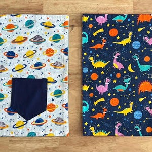 May include: Two fabric placemats with a space theme. One placemat is white with colorful planets and stars, the other is dark blue with colorful dinosaurs and stars. Both placemats have a pocket in the bottom right corner.
