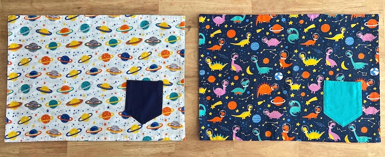 May include: Two fabric placemats with a space theme. One placemat is white with colorful planets and stars, the other is dark blue with colorful dinosaurs and stars. Both placemats have a pocket in the bottom right corner.