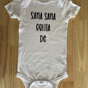 May include: White baby onesie with black text that reads "Sana Sana Colita De".
