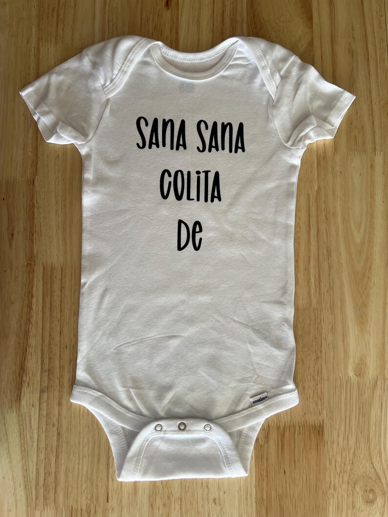 May include: White baby onesie with black text that reads "Sana Sana Colita De".
