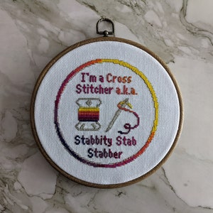 Pode incluir: Um aro de bordado branco com um desenho de ponto cruz que diz "I'm a Cross Stitcher a.k.a. Stabbitty Stab Stabber". O desenho apresenta um carretel de fio com uma agulha e fio.