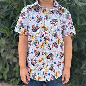 May include: A white short-sleeved button-down shirt with a repeating pattern of Mario Kart characters, including Mario, Luigi, and Toad. The shirt has a blue collar and blue buttons. The text "VROOM!" and "DRIFT!" appears on the shirt.