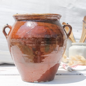 Puede incluir: Un gran tarro de cerámica esmaltada marrón con dos asas. El tarro tiene un aspecto rústico y vintage y es perfecto para almacenar alimentos u objetos decorativos.