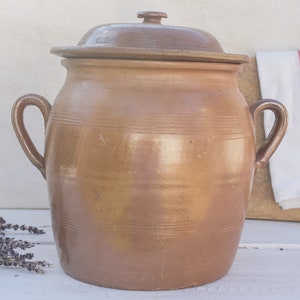 May include: A large, brown earthenware crock with a lid. The crock has a wide mouth and two handles. The crock is sitting on a white surface.