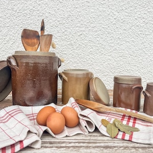 May include: A collection of brown ceramic crocks with lids, some with wooden handles, and wooden spoons. Four brown eggs are on a white and red checked cloth in front of the crocks.