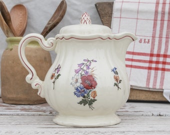 May include: A white ceramic teapot with a floral design featuring pink, red, and blue flowers. The teapot has a red rim and a red handle.