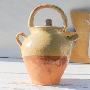 May include: A rustic, hand-crafted ceramic pitcher with a light yellow glaze and a brown base. The pitcher has a handle and a spout, and a small lid.
