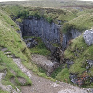 Peut inclure: Un paysage naturel avec une gorge profonde, des falaises rocheuses grises et des pentes herbeuses vertes. Un sentier mène dans la gorge, avec de l'herbe et du gravier au fond. Le ciel est couvert.