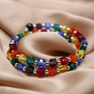 Puede incluir: Un brazalete de cuentas de colores arcoíris con separadores plateados. El brazalete está hecho con cuentas cuadradas de color rojo, naranja, amarillo, verde, azul, índigo y violeta.