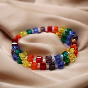 May include: A rainbow-colored beaded necklace with square-shaped beads. The necklace is made of red, orange, yellow, green, blue, indigo, and violet beads.
