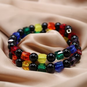May include: A rainbow-colored beaded bracelet with black beads and square-shaped beads. The bracelet is made of glass beads and is wrapped around a brown fabric background.