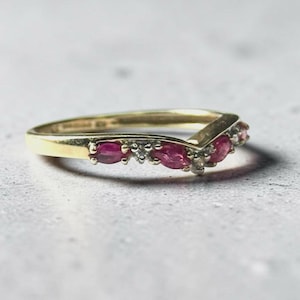 Könnte beinhalten: Ein V-förmiger Goldring mit einer Reihe von rubinroten Steinen im Marquise-Schliff, die sich mit kleinen klaren Akzentsteinen abwechseln. Die Ringschiene hat eine glatte, polierte Oberfläche und folgt einer Chevron-Silhouette mit Edelsteinen.
