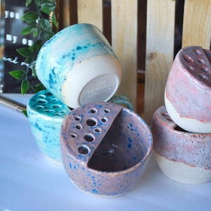 May include: A collection of handmade ceramic pieces in various shapes and sizes. The pottery features speckled glazes in shades of blue, pink, and purple, with white bases. Some pieces have holes, and one has a handle. The items are displayed on a white surface.