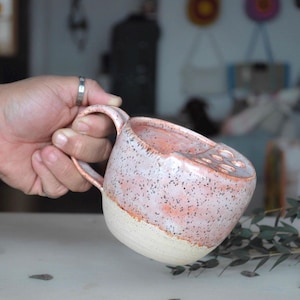 Op de afbeelding: Een handgemaakte keramische mok met een gespikkelde roze en witte buitenkant. De mok heeft een unieke vorm met een brede opening en een comfortabel handvat. De onderste helft is wit en de bovenste helft is roze met donkere spikkels.