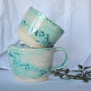 Puede incluir: Dos tazas de cerámica hechas a mano. Las tazas están apiladas, una encima de la otra. Las tazas tienen un esmalte moteado turquesa y blanco. Una taza tiene un asa. Las tazas están sobre una superficie blanca.
