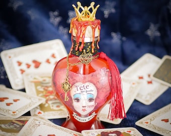 May include: A red glass bottle with a gold crown stopper and a red tassel. The bottle has a label that says "Tete qui gonfle" and "Drink Me". The bottle is surrounded by playing cards with hearts.