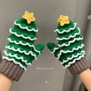 May include: A pair of green and white crocheted Christmas tree mittens with brown cuffs and yellow star embellishments.  The text "Let's Yarn Over Over" is visible in the background.