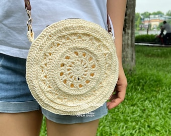 Puede incluir: Un bolso redondo de crochet blanco con una correa marrón. El bolso tiene un diseño circular con un círculo central y múltiples capas de detalles de crochet.