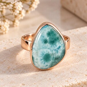 May include: A ring featuring a polished, teardrop-shaped larimar gemstone with swirling patterns of light blue and deep teal. The stone is set in a smooth, rose gold-toned metal band that encircles the finger with a simple, clean design.