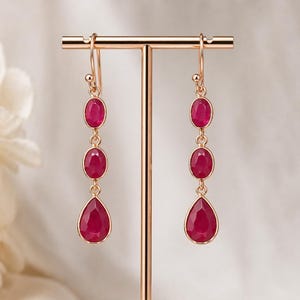 May include: A pair of drop earrings featuring three ruby-red gemstones in oval and teardrop shapes, set in gold-coloured metal. The earrings are displayed on a gold-coloured stand with a white marble base. The background is a soft, neutral colour.