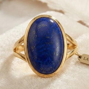Pode incluir: Um anel de ouro com uma grande pedra preciosa de lápis-lazúli de formato oval com manchas douradas naturais. A banda tem um design dividido nas laterais e uma pequena etiqueta retangular presa ao anel traz impresso o texto LAPIS LAZULI.