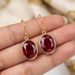 May include: A pair of gold-colored earrings with oval-shaped, deep red gemstones. The gemstones are set in a gold bezel and suspended from a simple gold hook. The earrings are displayed against a soft, neutral background.