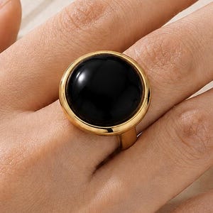 May include: A gold-toned ring featuring a large, smooth, circular black stone set in a polished metal bezel. The ring has a simple, solid band and a reflective surface on the black stone, showcasing a clean, rounded design for the finger.
