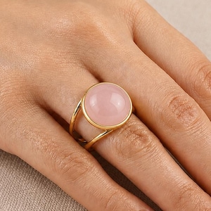 Op de afbeelding: Een goudkleurige ring met een gladde, ronde, lichtroze cabochonsteen. De ringband bestaat uit twee dunne, parallelle goudkleurige draden die samenkomen bij de zetting, wat zorgt voor een open bypass-ontwerp rondom de vinger.