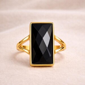Könnte beinhalten: Ein goldfarbener Ring mit einem großen, rechteckigen schwarzen Edelstein. Der Edelstein ist facettiert und reflektiert das Licht. Das Ringband hat ein geteiltes Design. Ein Schmuckstück.