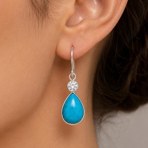May include: Silver drop earrings with turquoise teardrop stones. Each earring has a bright turquoise, teardrop-shaped stone set in silver. Above the turquoise is a small, round, clear gemstone. The earrings hang from silver hooks.