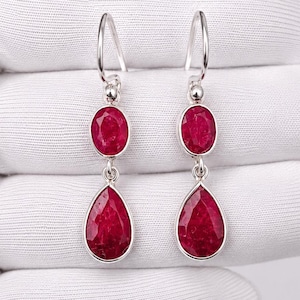 Può includere: Un paio di orecchini pendenti in argento con pietre preziose rosso scuro di forma ovale e a goccia. Gli orecchini sono presentati su uno sfondo bianco, evidenziando il colore vibrante e il design dei gioielli.