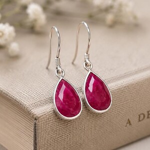 Puede incluir: Pendientes colgantes de plata con piedras preciosas rojo rubí facetadas en forma de lágrima. Los pendientes se muestran sobre un libro. Flores blancas delicadas en el fondo. Las piedras miden aproximadamente 1,5 cm.