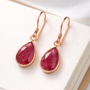 May include: A pair of rose gold teardrop earrings with faceted, deep red gemstones. The earrings are displayed on a white surface, showcasing their elegant design and color.