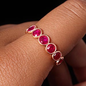 May include: A gold ring featuring a series of vibrant red gemstones. The gemstones are oval-shaped and set closely together, creating a continuous band of color. The ring is displayed on a finger against a dark background.