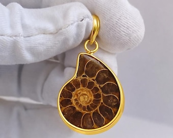 May include: A gold-plated pendant featuring a polished, fossilized ammonite. The pendant showcases the intricate spiral pattern of the ancient sea creature, set within a gold frame. The pendant is suspended from a gold bail.